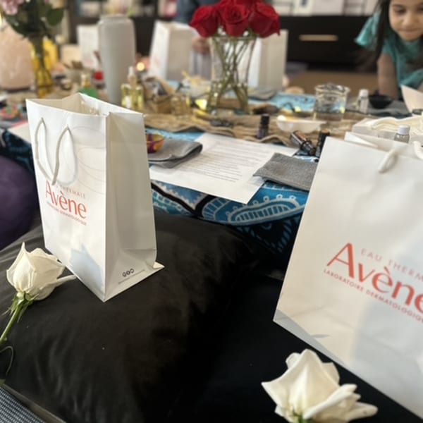 Two white Avene shopping bags with white roses, a table with red roses, and a child in the background.