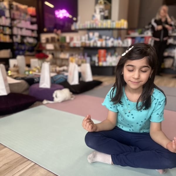 Young girl meditating on a yoga mat with eyes closed, surrounded by pillows and gift bags.